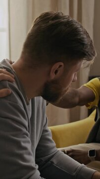Vertical shot of sad man sitting on sofa, taking sip of whiskey while his best friends patting his shoulder, trying to support him