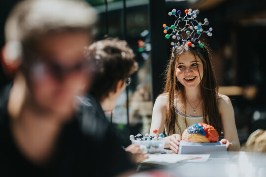 A young woman smiles while holding a colorful brain model and wearing a molecular structure headpiece. Friends sit around a table studying science together in a casual outdoor setting.