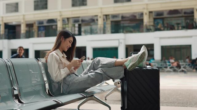 Portrait of happy Asian lesbian texting on smartphone at train station. Young trans woman puts legs onto luggage scrolling social media in waiting hall side view. Ready for journey