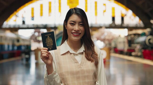 Portrait of cheerful Asian transwoman showing passport standing on railway platform. Positive young lesbian traveler with personal ID waits for train at station. Business journey