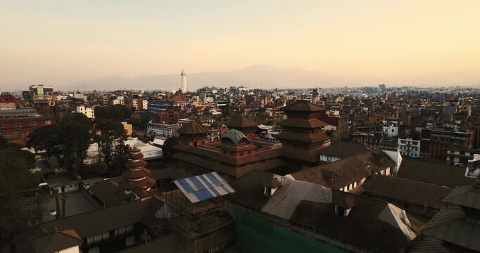 Atmospheric aerial drone shot of Basantapur Durbar Square's ancient pagoda rooftops under a soft pastel sky, with the Dharahara tower visible in the distance.