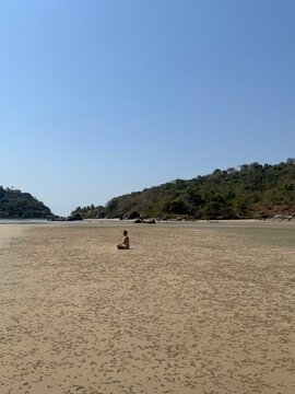 Minimalist scene of a woman in a swimsuit meditates or sits alone on the vast sand of Palolem Beach, Goa, India. Wellness and mindfulness concept during travel.