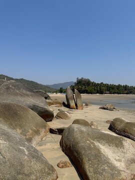 Large volcanic rock formations on the sandy shore of Palolem Beach in South Goa, India. Tropical coastline with palm trees under a clear blue sky.