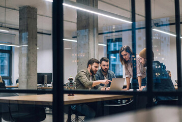 Obraz premium Creative startup team collaborating on a laptop in a modern glass office with a robot toy on the desk, viewed through a partition
