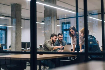 Creative startup team collaborating on a laptop in a modern glass office with a robot toy on the desk, viewed through a partition © IBEX.Media