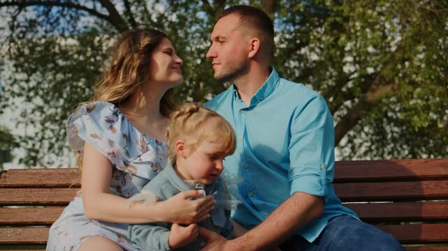 Family of three sits on park bench. Mother wears floral dress, father wears blue shirt. Child rests head on mother's shoulder. They smile and look at each other. Scene shows affection and calm