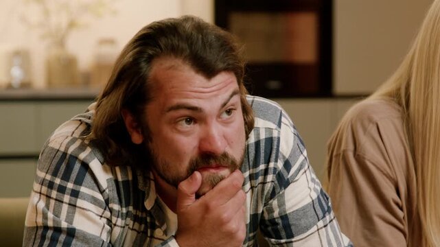 Caucasian male with beard wearing plaid shirt gestures while engaged in conversation with a woman in a cozy indoor setting, modern kitchen visible in the background