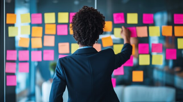 Businesswoman in dark suit arranging colorful sticky notes on glass wall in modern office, collaborative workspace with blurred furniture in the background