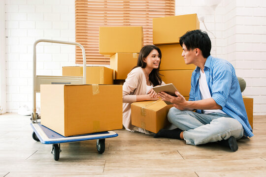 Couple sitting on the floor among cardboard boxes, using a tablet and planning a home move with a loaded dolly in a bright living room