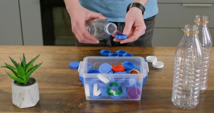 Adult man twists blue cap from plastic bottle near sorting container on table. Person drops loose lid into box continuing separating bottle parts for recycling