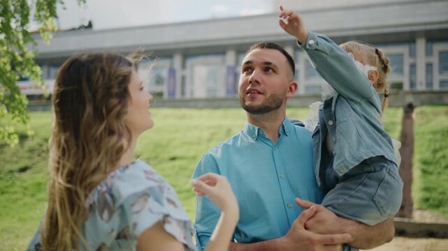 Happy family of three smiles outdoors near modern building. Mom laughs, dad holds toddler who points upward. Scene shows joyful bonding in green space. Suitable for parenting, family