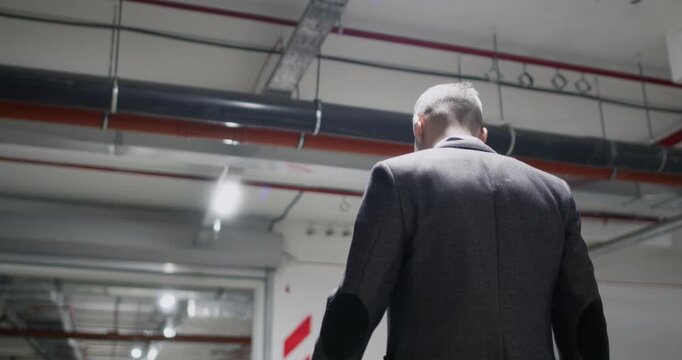 Serious businessman walks through underground parking area of residential building. Man observes structural elements and parked vehicles during detailed inspection