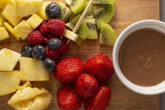 Fresh fruit platter with chocolate dip and skewers on wooden board, strawberries, pineapple chunks, kiwi slices, blueberries
