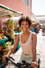 Fototapeta premium African American woman smiling while riding a bicycle at an outdoor farmers market. Urban lifestyle and summer outdoor recreation. Healthy living and leisure activity in city center.