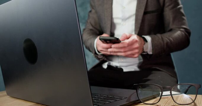 Male scammer uses smartphone while sitting near laptop during online fraud process. Man types on phone skreen managing communication for deceptive digital scheme