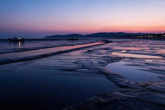 Twilight paints the sky in soft hues over a tidal flat with calm water. Fishing boats rest on wet sand near a distant coastline with town lights. Mud patterns reflect the colorful dawn light.