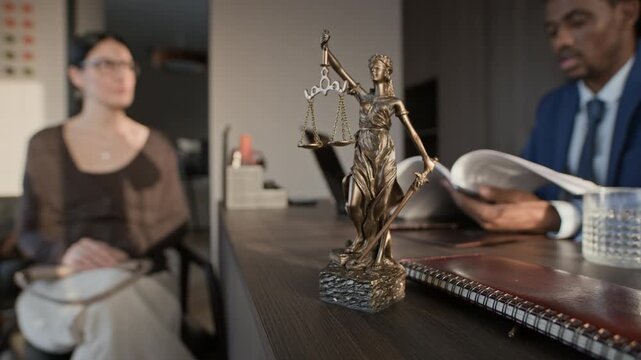 Close up of Themis statue on wooden table, African American lawyer chatting with clients, discussing settlement agreement