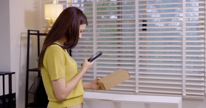 Online seller confirming product shipment on smartphone, scanning barcode on parcel, managing order details in home office, small e-commerce business preparing inventory for delivery.