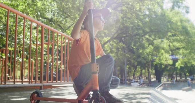 Boy squats near scooter as holding handlebar during break in skate park. Child rests on curb while studying surroundings and keeping orange ride steady nearby