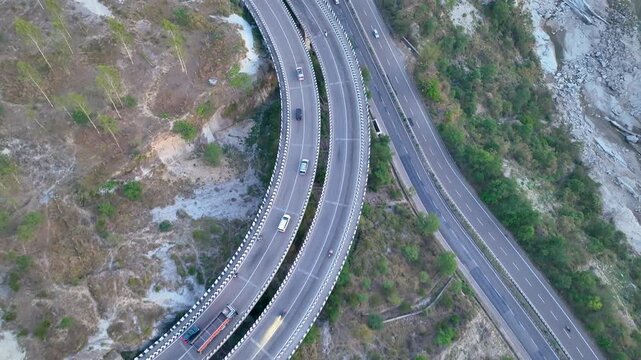 Top-down aerial view of curved road infrastructure. Drone footage capturing transport trucks and commuter vehicles navigating a sweeping curve on a major national highway in India. 