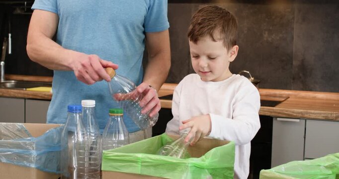 Boy drops empty plastic bottle into green recycling bin in kitchen. Father stands nearby guiding sorting while preschooler child places container with careful hands