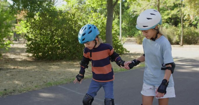 Girl teaches younger brother to skate as boy loses balance on park path. Sister holds hand steadying child while inline rollerblades slide forward carefully nearby