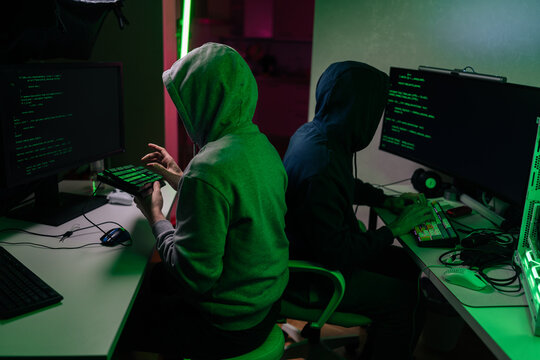 Two anonymous hackers in hoodies collaborating on cybercrime. Programmers work late at night in dark room with green light. Concept of cybersecurity, data theft, teamwork in illegal activity.