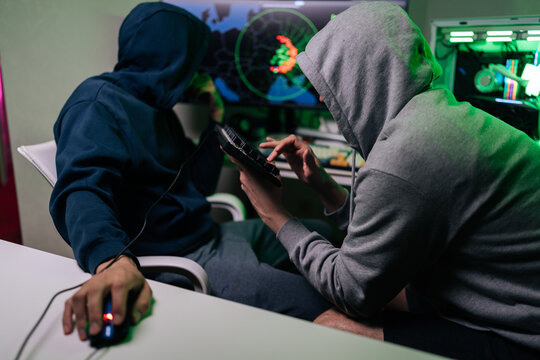 Two unrecognizable people in hooded sweatshirts sit at desk in dark room. They work on computer, focused on screen with world map and code. Green light creates mysterious, tense atmosphere.