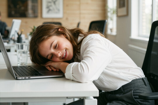 A hopeful young girl sleeps with her head down on a modern office desk beside a laptop and glass of water, exuding quiet optimism.