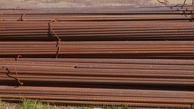 Large bundles of rusty steel reinforcing bars, known as rebar, are stacked horizontally on the ground, ready for use in reinforced concrete construction at an industrial building site