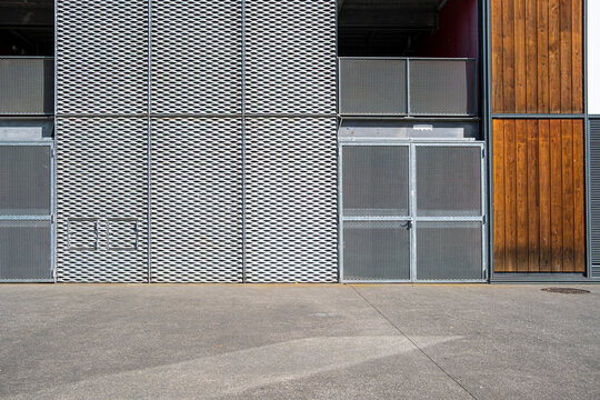 Metal mesh doors sit beside warm wood panels on a quiet urban sidewalk, giving the industrial facade a layered exterior texture along the street