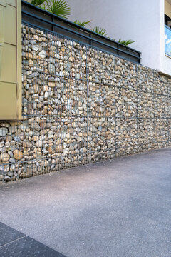 A narrow urban walkway follows a gabion wall, pulling perspective along stone texture and pavement beside a clean facade exterior