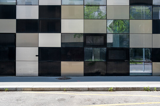 A geometric glass facade holds a grid of dark and pale panels above the sidewalk, where street reflection turns the urban exterior into a calm pattern