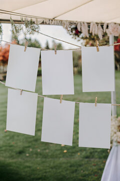 Outdoor wedding seating chart mockup with six hanging vertical cards on rope. Blank table plan template with wooden clothespins on blurred green lawn background.
