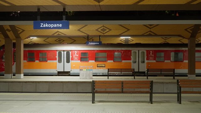 Red passenger train stands at empty station platform at night in Zakopane, Poland. Deserted railway terminal for travel to Tatra mountains during evening commute and night transit.