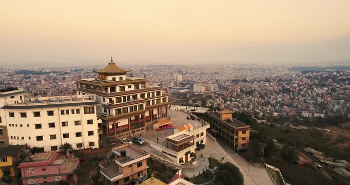 Aerial drone shot of the Chovar Gumba monastery in Nepal perched above the vast Kathmandu cityscape, with its pagoda spire glowing against a hazy golden dusk sky.