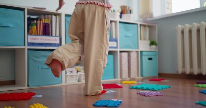Children balance barefoot on colorful textured sensory mats during a playful indoor activity in a bright classroom with storage cubbies