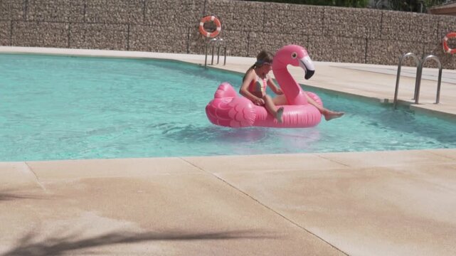 Little girl slowly floating away from the pool edge on a pink inflatable flamingo. Slow motion sstkSummer lifestyle video showing childhood vacation, pool recreation, sunny summer fun and playful