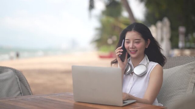 Asian happy digital nomad working on laptop at tropical beach, Beautiful freelance woman with headphones enjoying remote work and summer vacation, Flexible workation and travel lifestyle concept