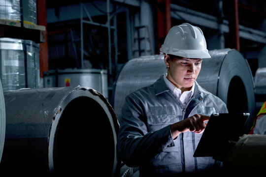 Young worker with tablet inspecting large metal tubes at factory during night shift. Engineer with helmet and safety vest working in industrial site. Inspector checking quality of product in a dark