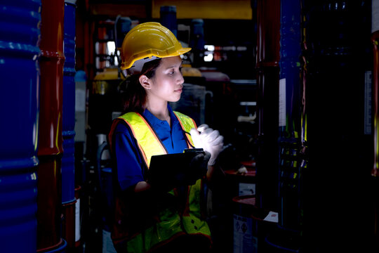 Asian female worker with tablet inspect product in large steel drums at factory during night shift. Engineer woman with helmet and safety vest work in industrial site, check product quality in a dark