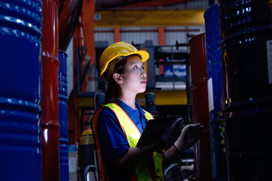 Asian female worker with tablet inspect product in large steel drums at factory during night shift. Engineer woman with helmet and safety vest work in industrial site, check product quality in a dark
