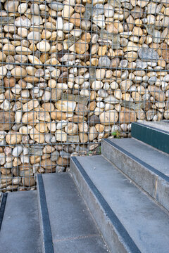 Concrete stairs meet a gabion wall where stone texture, handrail, pavement, and urban exterior details turn a small architectural corner into quiet geometry