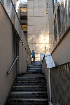 A lone person climbs concrete stairs through a narrow passage, where handrail, shadows, facade, and urban architecture compress the exterior depth