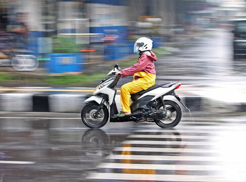 Side view of a person in a raincoat riding a scooter on a wet city street, panning shot
