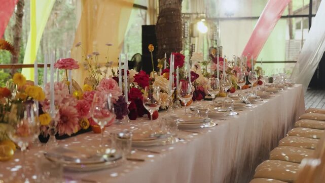 Luxury wedding banquet table setting with flowers and candles under outdoor tent in 4K slow motion. Beautifully decorated festive dinner table with floral centerpieces, wine glasses, and plates for a