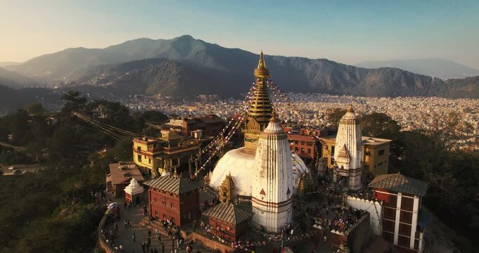 Aerial view of Swayambhunath stupa with its golden spire and colorful prayer flags glowing in warm sunset light, framed by misty Himalayan foothills.