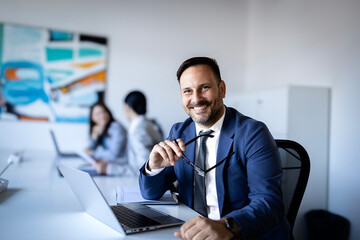Obraz premium Confident businessman smiling, holding glasses, working with laptop in modern office, colleagues in background
