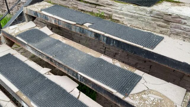 Worn treads on concrete steps showing torn coverings, rusted metal edges, gravel grit and moss between risers outdoor pickup point