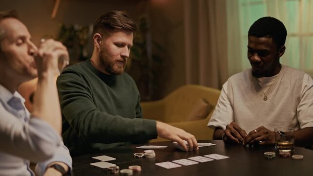 Handheld shot of biracial friends sitting at round table, drinking bourbon, chatting and playing poker